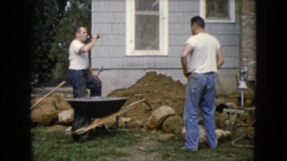 A man drinks from a brown bottle while another faces the work area near a wheelbarrow and dirt pile outside the gray house.