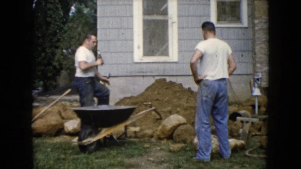 The final frame shows two men beside a wheelbarrow, rocks, and soil along the house wall during a backyard landscaping project.
