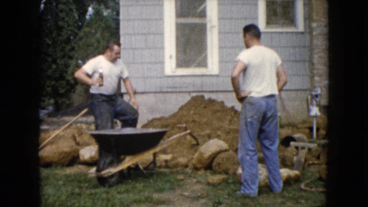 Two men stand near a wheelbarrow, soil pile, rocks, and a gray house exterior.