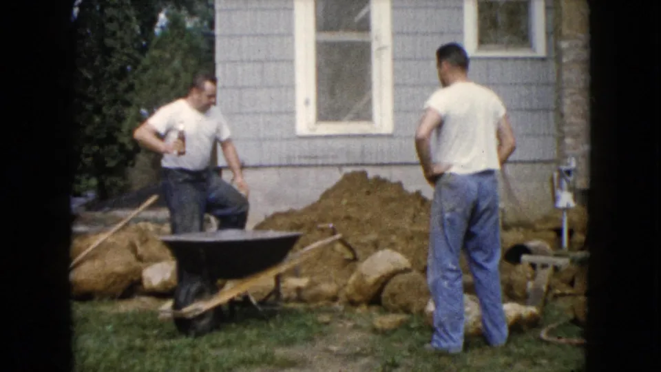 In Wauconda, Illinois, 1960, two men stand by a wheelbarrow and dirt pile during a residential lawn project.