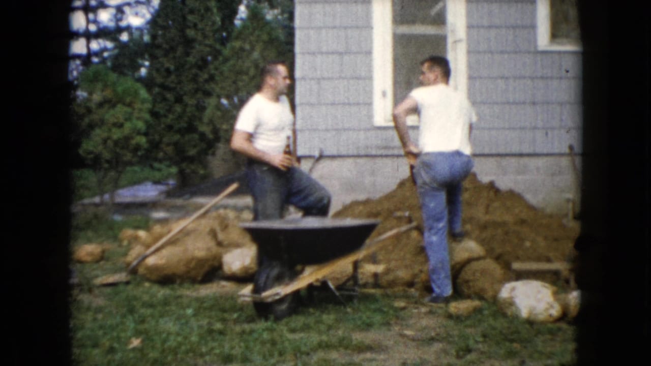 In Wauconda, Illinois in 1960, two adult men pause with shovels by a wheelbarrow during backyard digging near a house exterior.