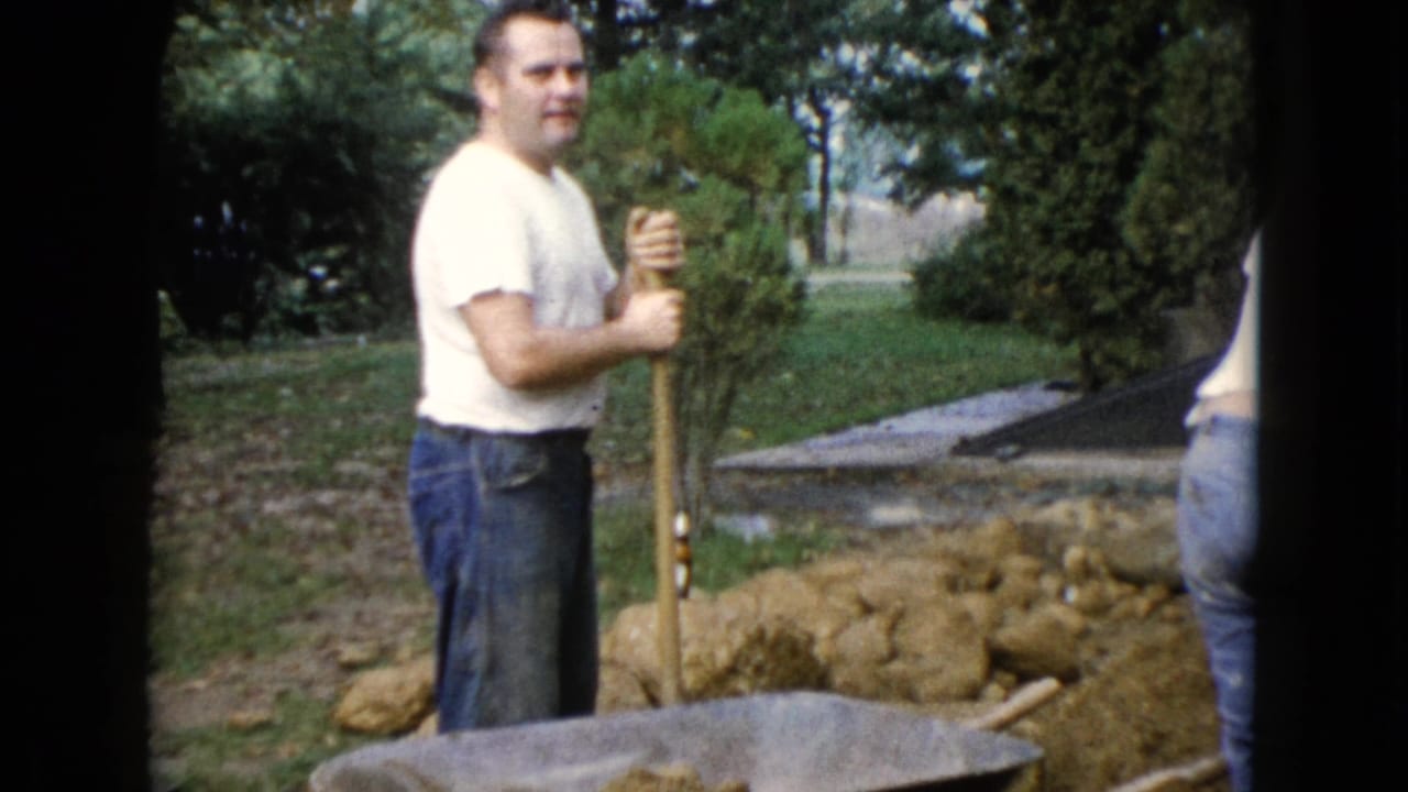 A man holds a shovel upright near a wheelbarrow, dirt piles, grass, and shrubs.