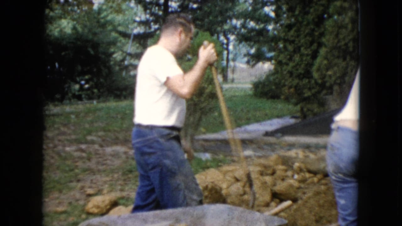 Man in a white T-shirt digs soil with a shovel beside a yard and house.