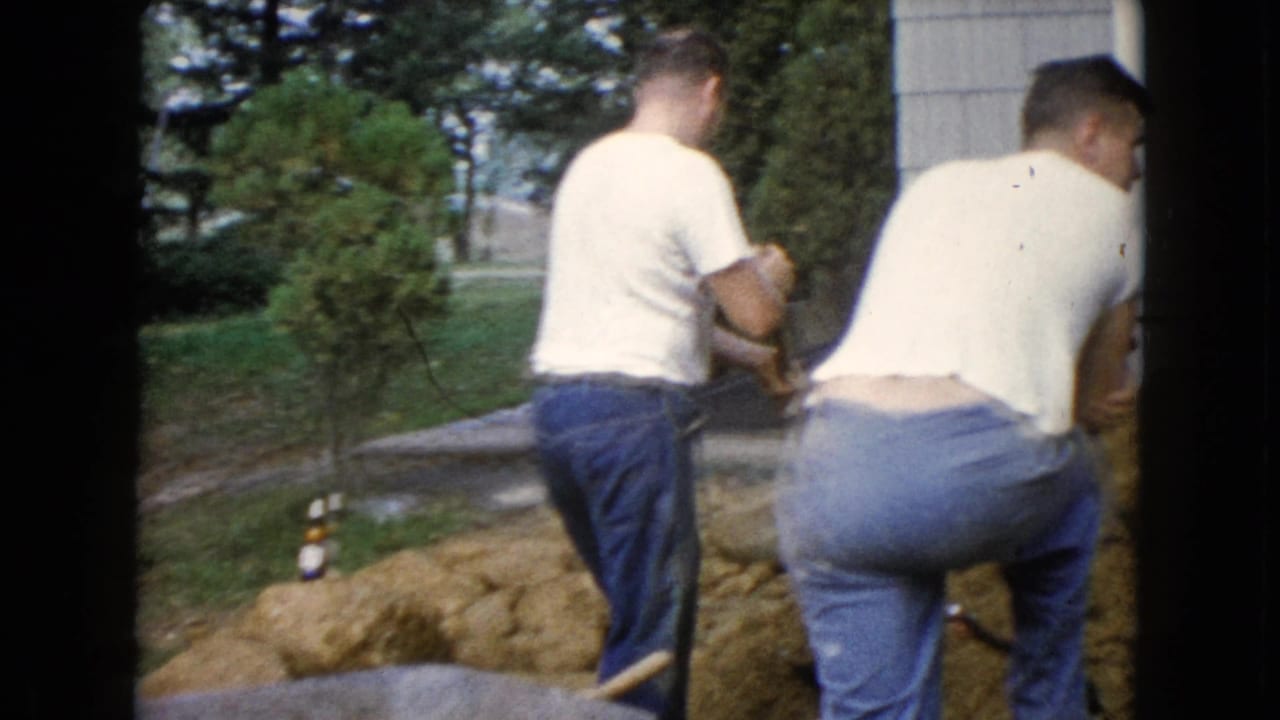 Two adult men in white T-shirts and jeans work beside a soil mound in a Wauconda yard, with trees and a house exterior behind them.