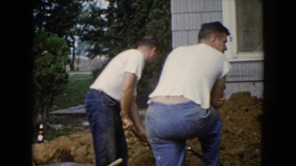 The men continue digging and moving soil beside a house exterior in Wauconda, with a lawn, trees, and scattered rocks in view.