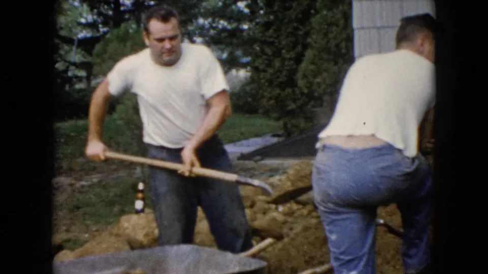 A man with a shovel stands near a wheelbarrow as another man works at the soil pile in the Wauconda backyard scene.