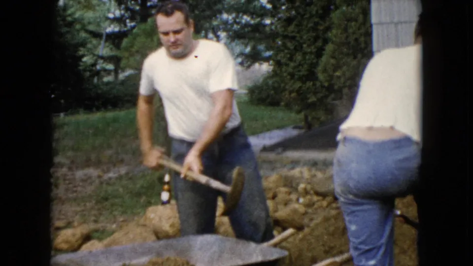In Wauconda, Illinois, 1960 home-movie footage, two adult men dig a trench beside a house with shovels and a wheelbarrow nearby.