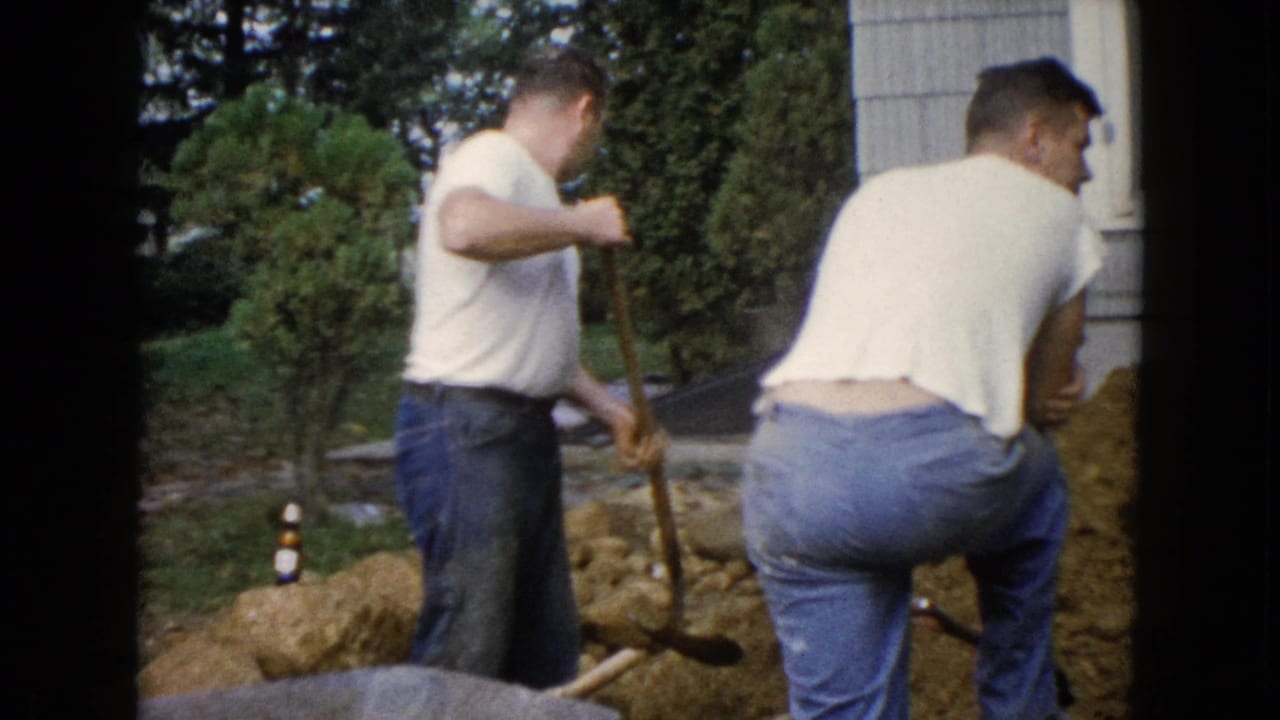 Two men stand near a dirt trench beside a house, each holding or using a shovel.