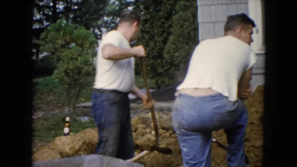 Two adult men continue trench work beside a house in Wauconda, Illinois, with shovels, dirt piles, shrubs, and lawn visible.