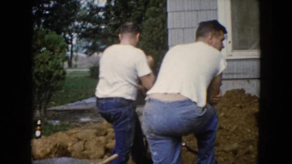 In this 1960 8mm frame, two men are seen from behind while working at a dirt trench beside a Wauconda house.