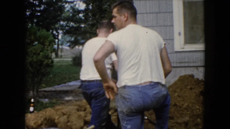 Two adult men stand near the trench beside a Wauconda, Illinois house, with lawn, trees, dirt, and a shovel handle visible.