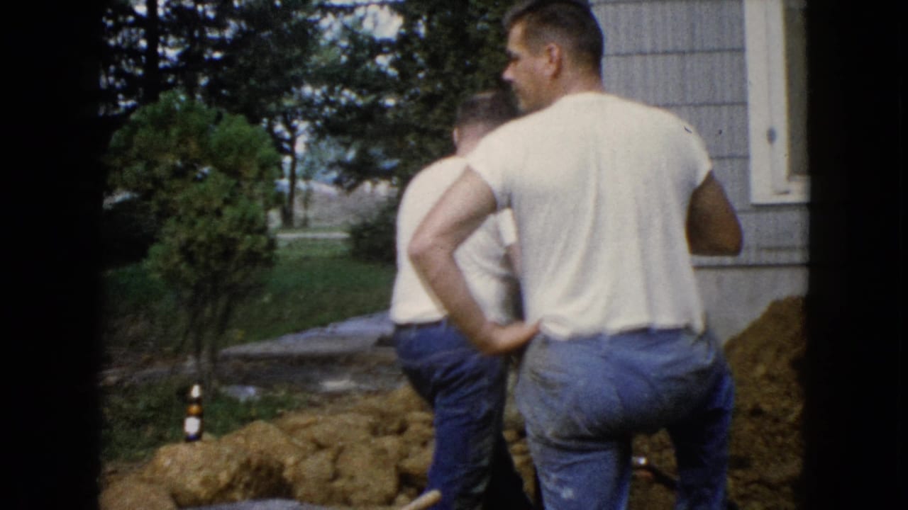 In Wauconda, Illinois, 1960, two adult men in white T-shirts and jeans stand near a dirt pile beside a suburban house.
