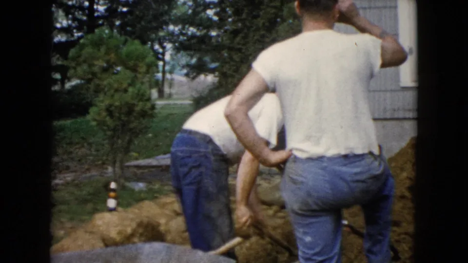 A 1960 Wauconda yard-work moment shows one man bending toward the soil while another stands nearby beside the house.