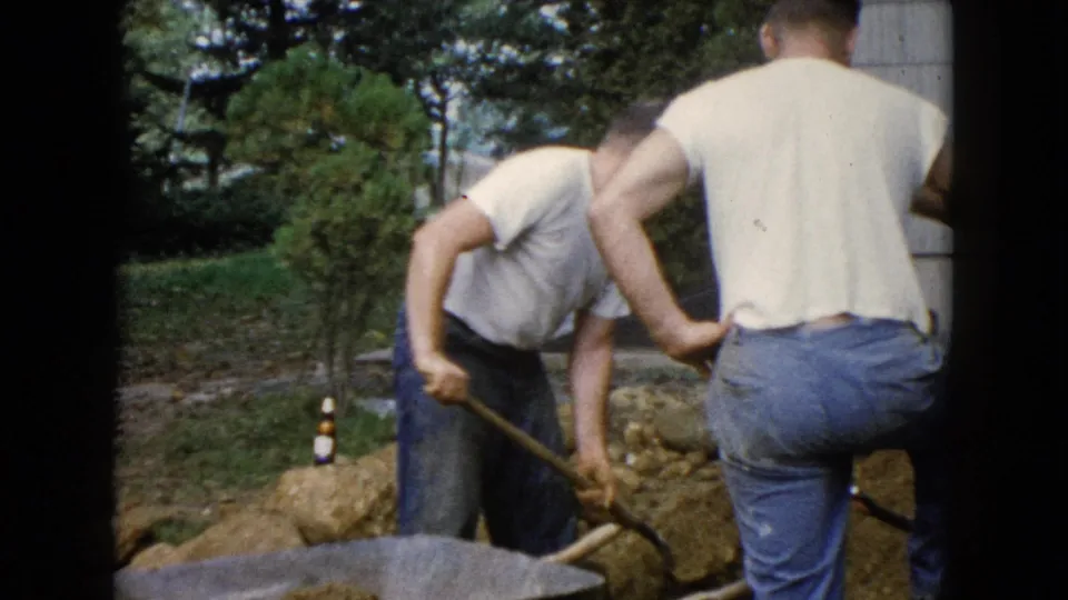 In the 1960 Wauconda home-movie scene, one man shovels dirt or rocks toward a wheelbarrow as another stands nearby.