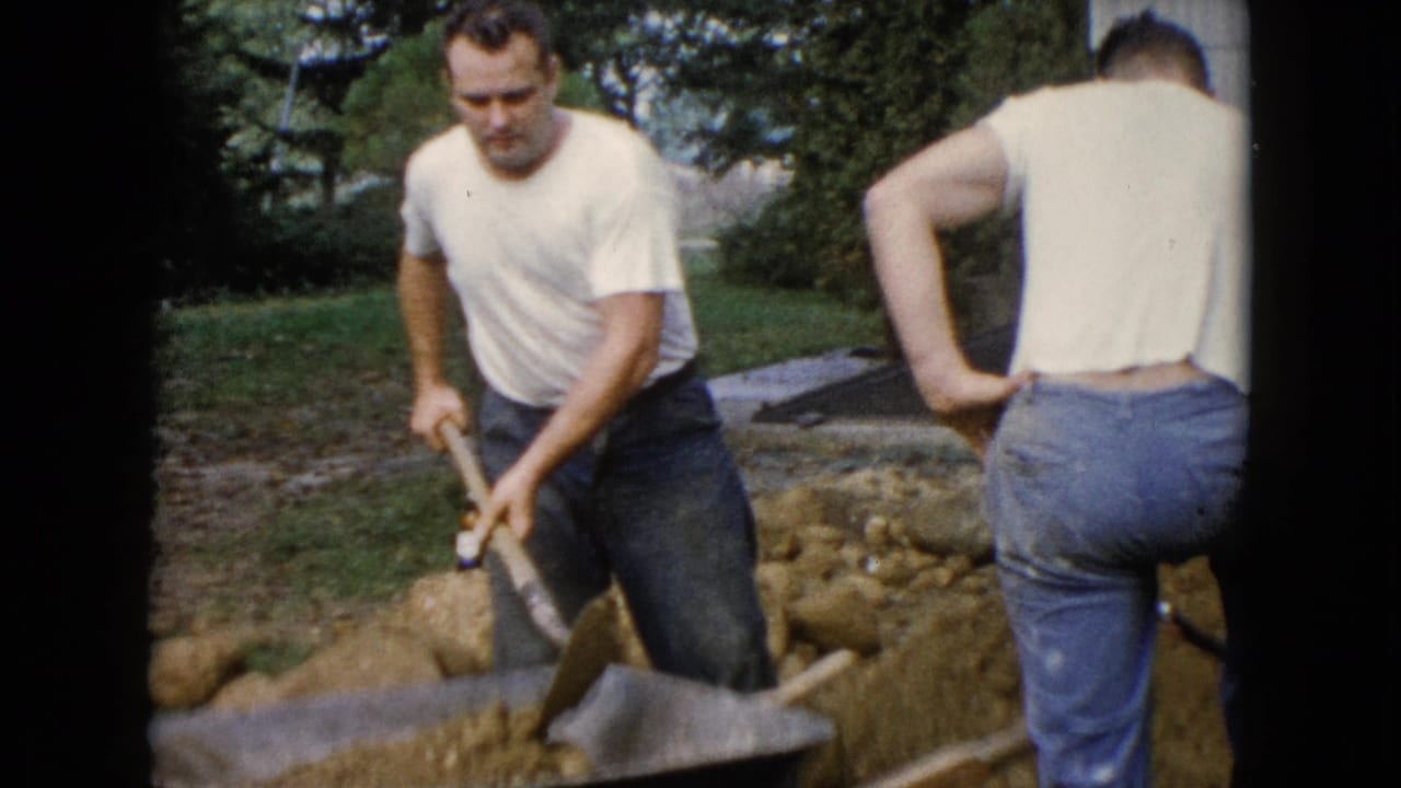Two adult men in a backyard, one shoveling dirt beside a wheelbarrow.