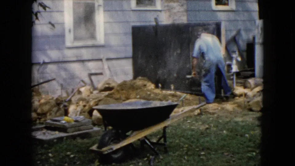 1960 Wauconda 8mm frame showing backyard work beside a house, with tools, soil, rocks, and a wheelbarrow.