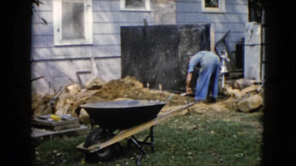 1960 Wauconda backyard construction frame with a man bending near a house and wheelbarrow in the foreground.