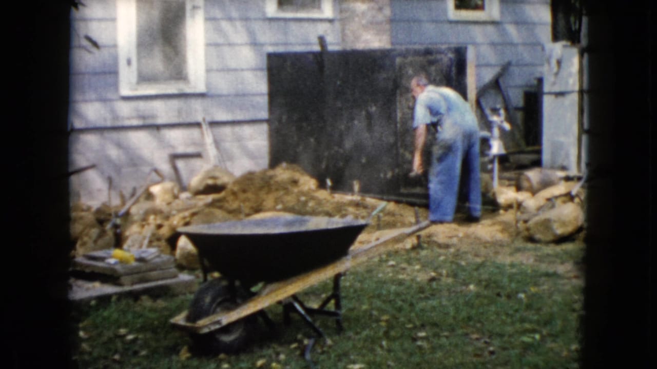 The man stands by a dirt mound and dark board beside the light-sided house during the Wauconda yard work scene.
