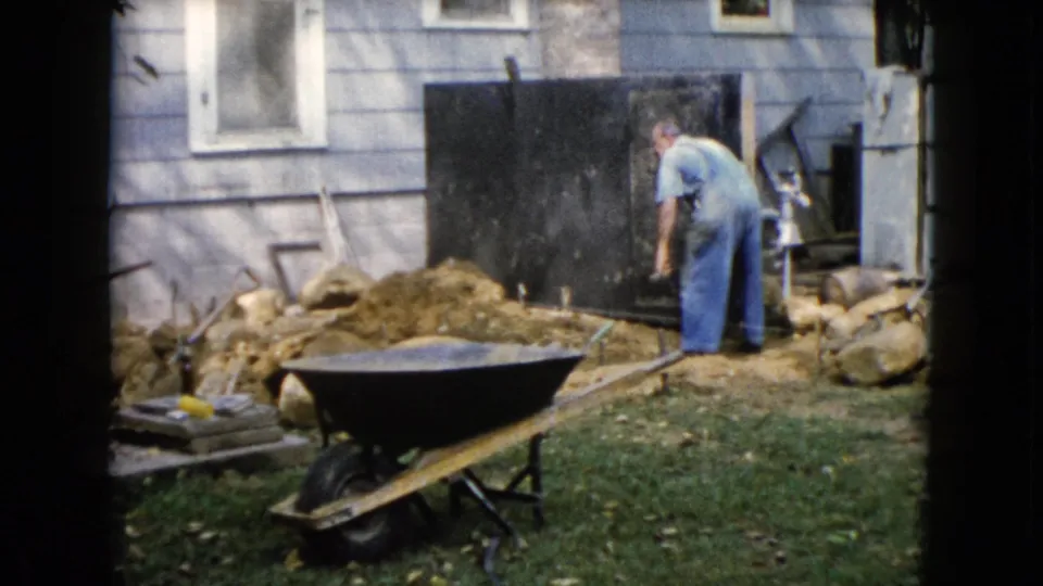 The man stands by a dirt mound and dark board beside the light-sided house during the Wauconda yard work scene.