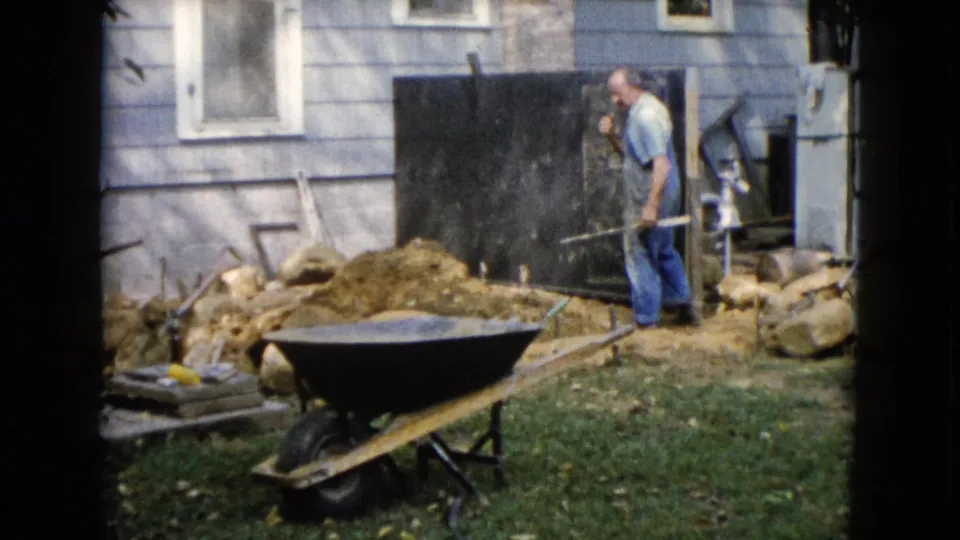 A man in work clothes holds a long-handled tool in a 1960 Wauconda backyard, with a wheelbarrow and house exterior nearby.