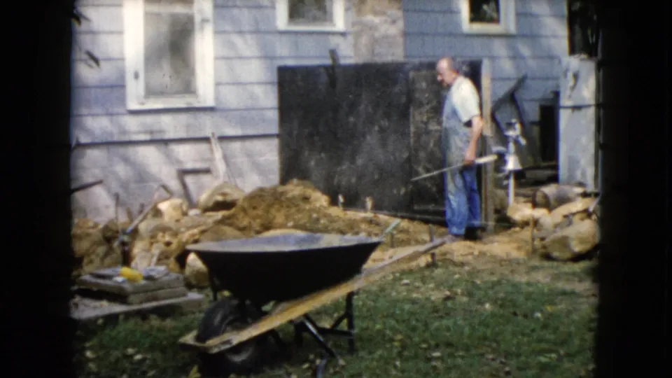 The backyard worker looks across the yard with a tool in hand, framed by the Wauconda house exterior and wheelbarrow.
