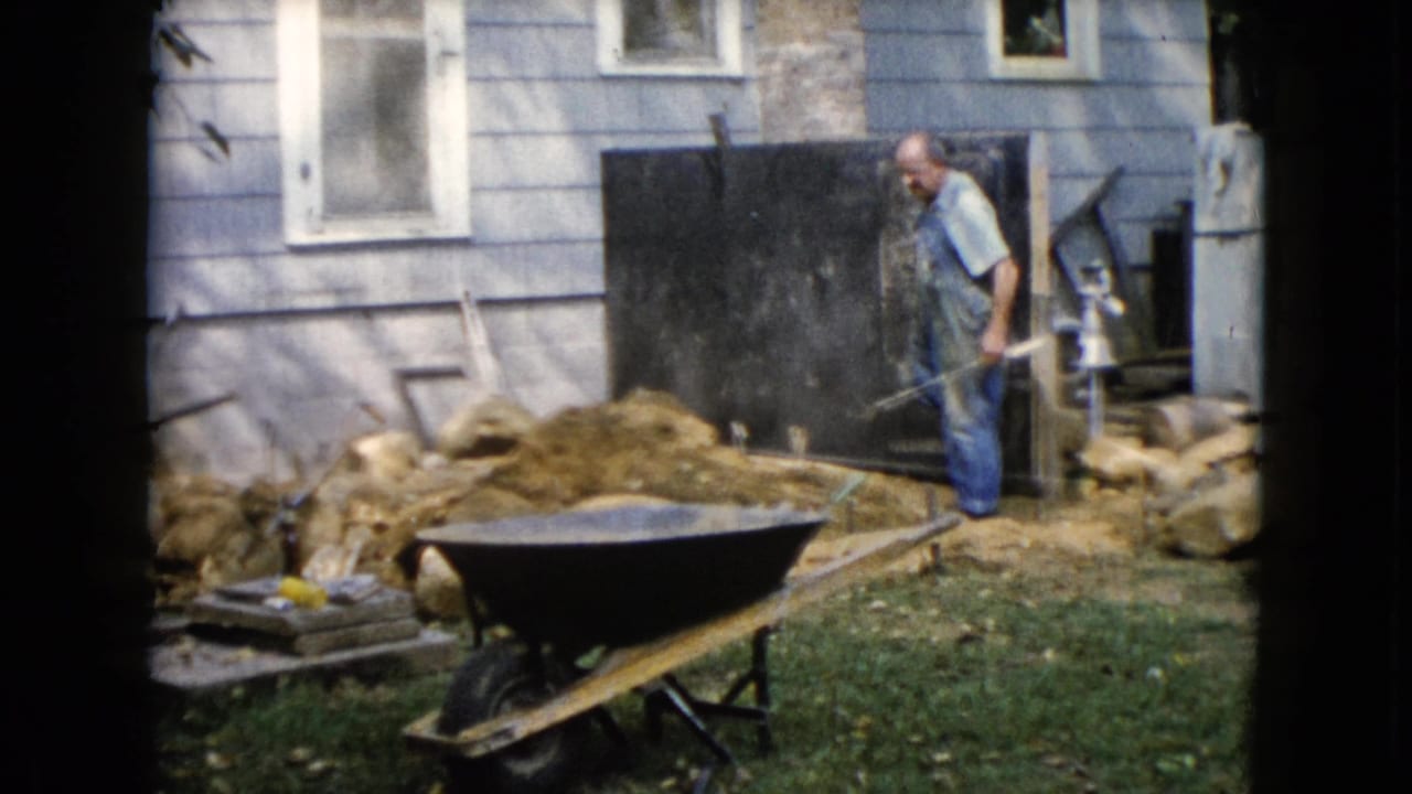 Man in blue overalls shoveling near a dirt pile and wheelbarrow beside a house.