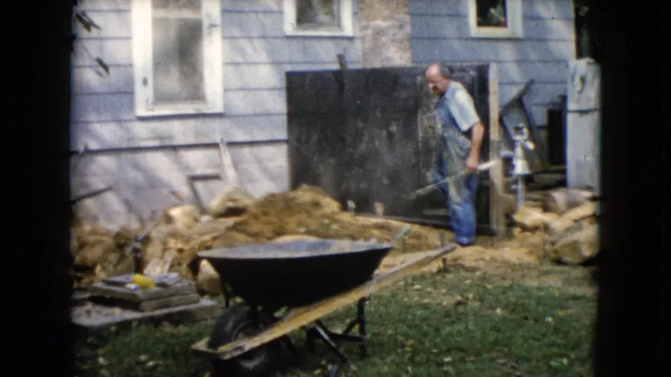 1960 Wauconda home-movie frame of a man in blue overalls working with a shovel near dirt, rocks, and a wheelbarrow beside a light blue house.