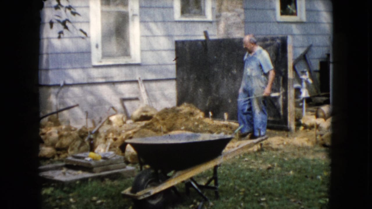 1960 Wauconda backyard frame showing a man holding a shovel beside a dirt pile, wheelbarrow, rocks, and the side of a light blue house.