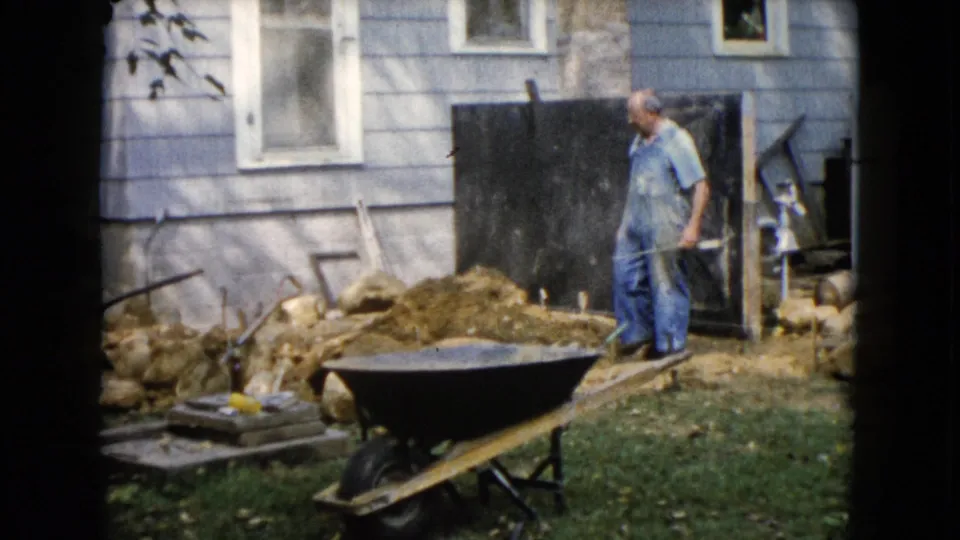1960 Wauconda backyard frame showing a man holding a shovel beside a dirt pile, wheelbarrow, rocks, and the side of a light blue house.