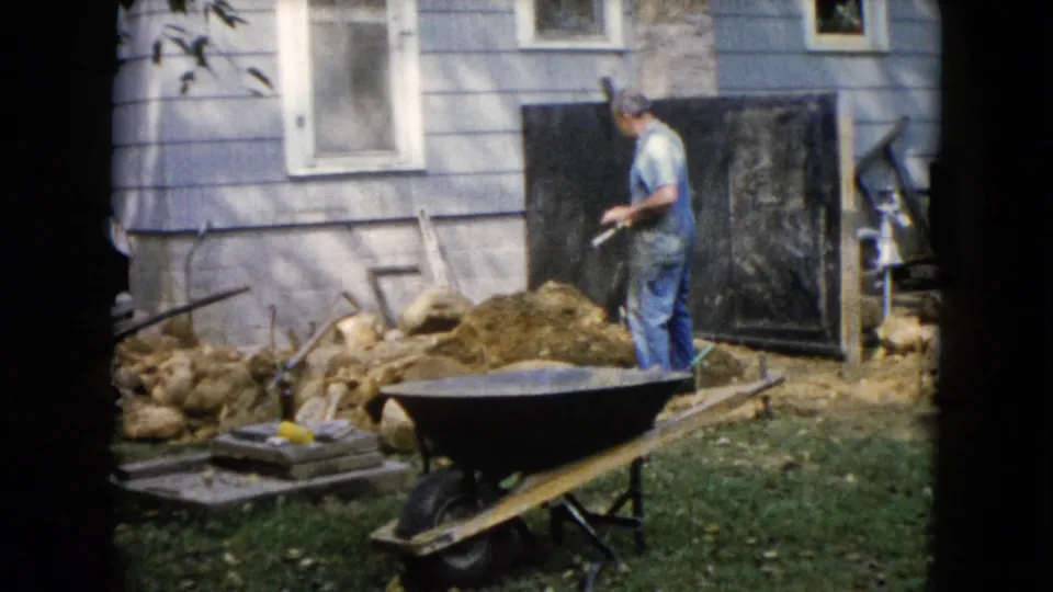 1960 Wauconda frame of a man in overalls working close to the house wall, with a wheelbarrow, dirt, rocks, and tools in the backyard.