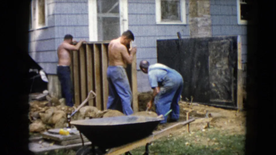 1960 Wauconda backyard frame showing three men, two shirtless, working around a wooden frame near dirt, rocks, and a wheelbarrow by the house.