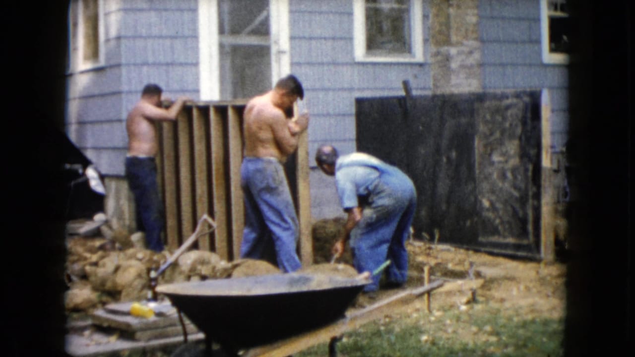 Three men work beside a wooden wall frame behind a blue-sided house with a wheelbarrow in front.