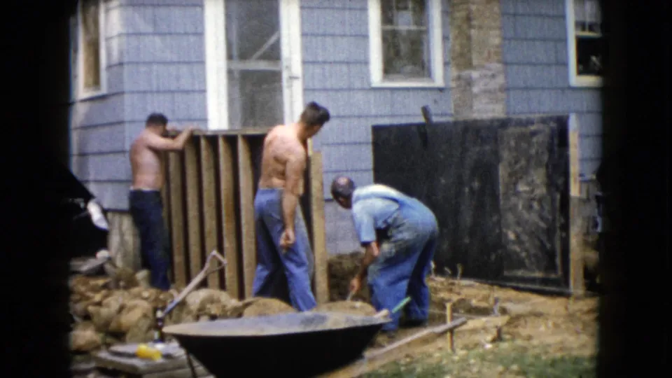 1960 Wauconda, Illinois frame showing men continuing work on a wooden wall frame behind a blue-sided house.
