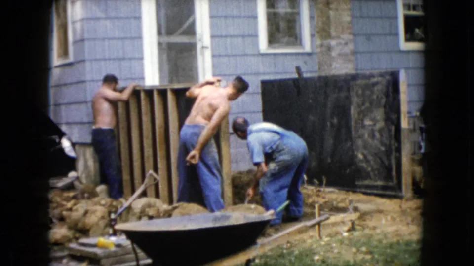 1960 Wauconda home-movie frame with one man stretching or leaning back during backyard wall construction.