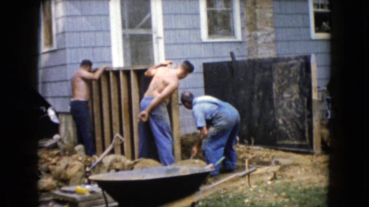 Three men work beside a blue-shingled house with wood framing, rocks, tools, and a wheelbarrow nearby.