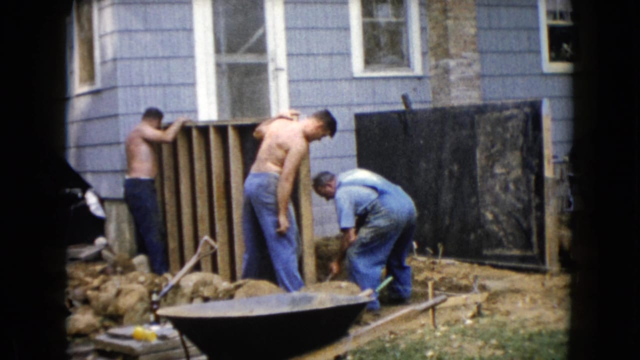 Three men continue backyard construction work beside a blue house in Wauconda, with a wheelbarrow and loose rocks in view.