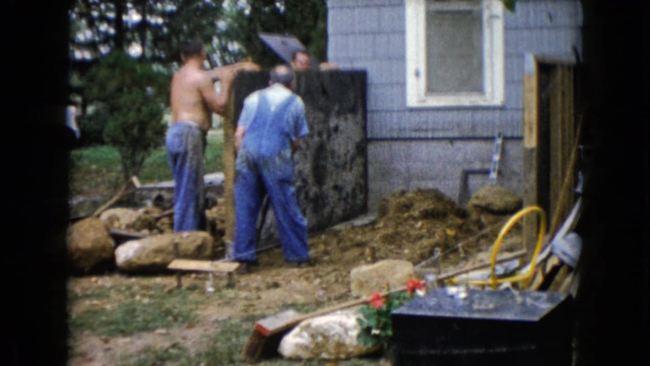 In supplied 1960 Wauconda home-movie footage, three men work outside a gray-sided house with rocks, boards, and yard tools nearby.