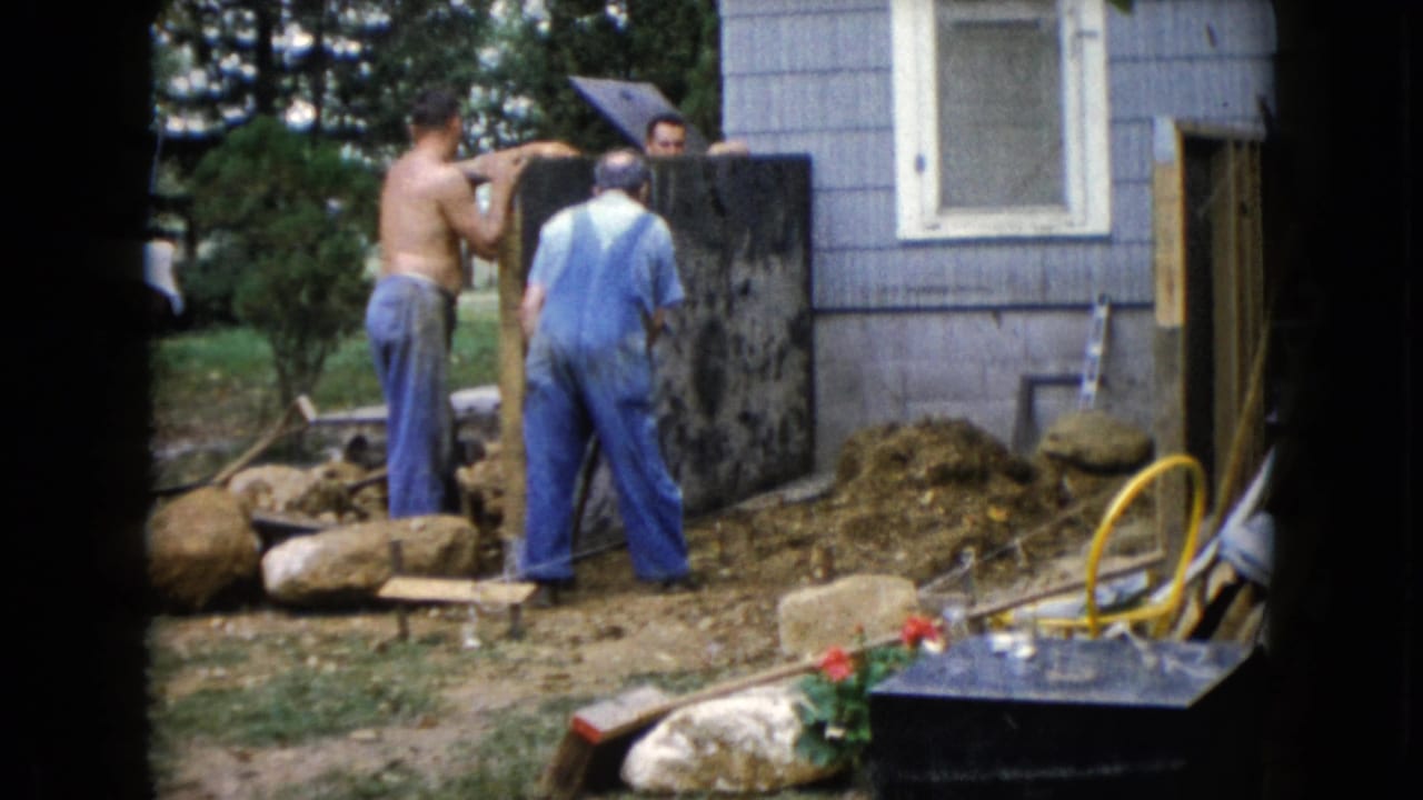 Three men continue work beside a gray-sided house in Wauconda, with a dark construction panel, soil, rocks, and hose visible.