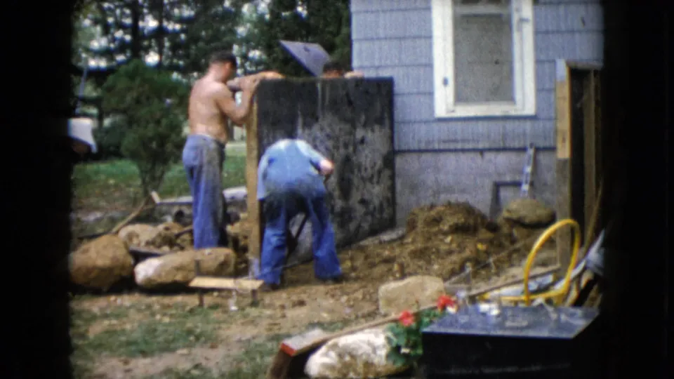 A man in blue overalls bends near the base of a dark panel as another man stands by the gray house in the 1960 Wauconda footage.