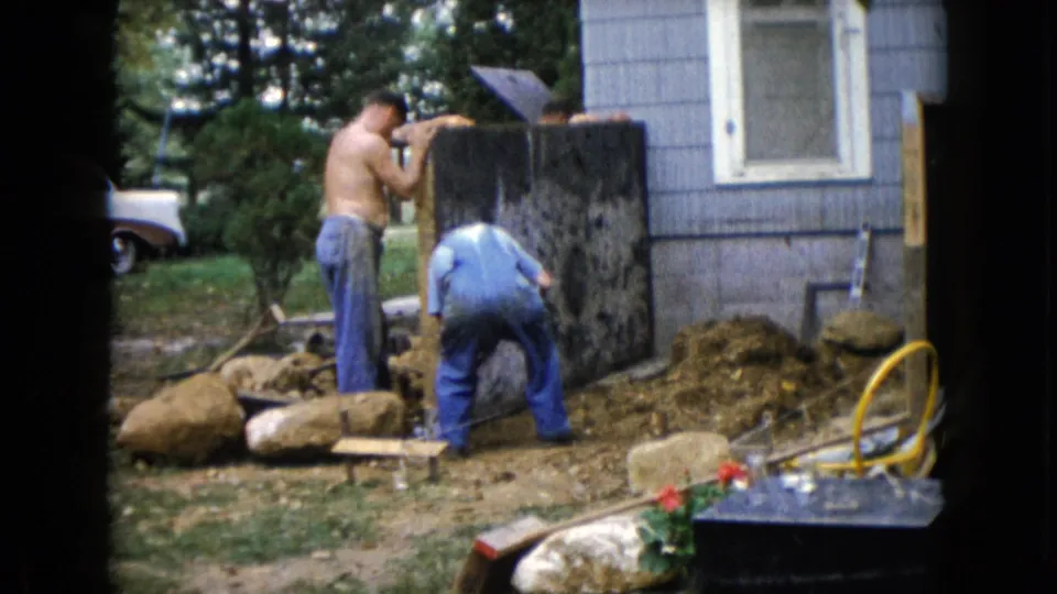 Two men remain visible beside the dark panel outside the gray-sided house, surrounded by dirt, rocks, boards, and a yellow hose.
