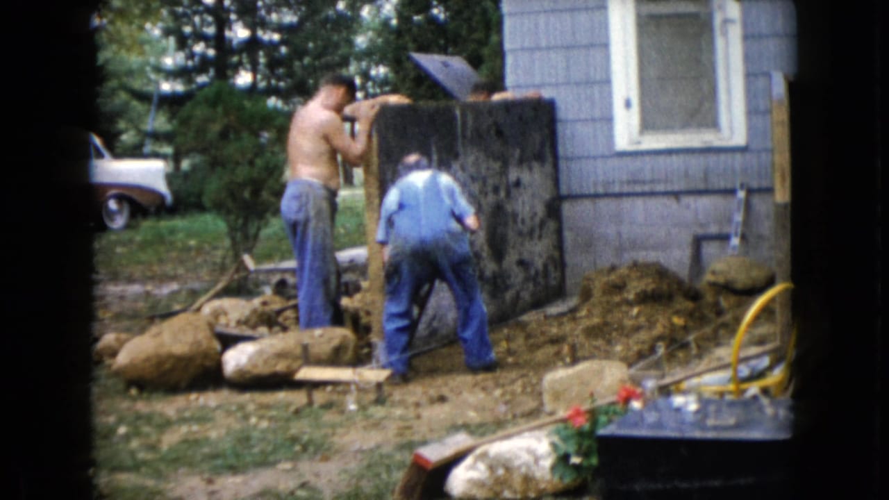 In 1960 Wauconda, Illinois home-movie footage, two men work near a dark upright slab beside a blue-sided house and yard debris.