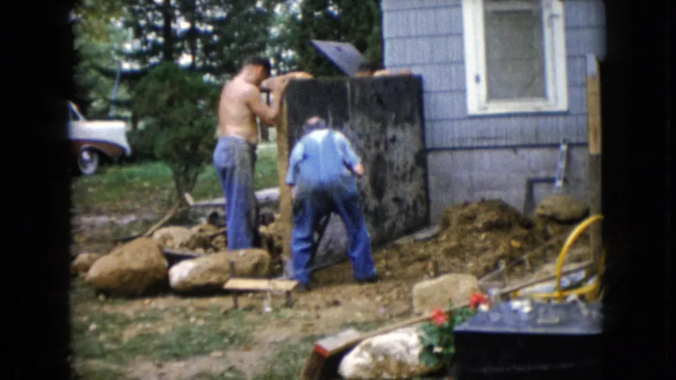 In 1960 Wauconda, Illinois home-movie footage, two men work near a dark upright slab beside a blue-sided house and yard debris.
