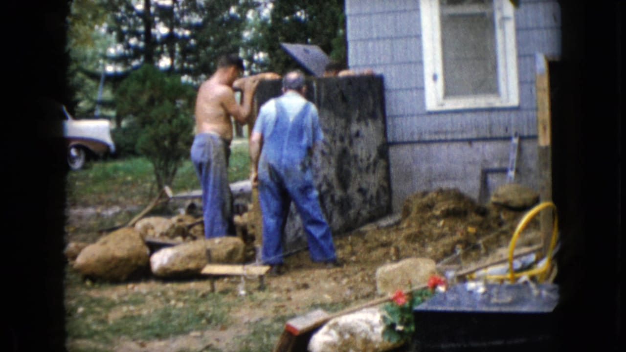 A shirtless man and a man in overalls stand beside a dark slab near a house.