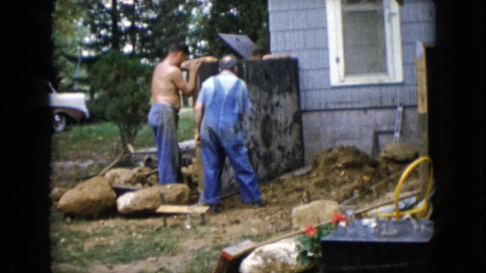 Two men, one shirtless and one in blue overalls, stand close to a dark slab outside a blue-sided house in Wauconda, Illinois.