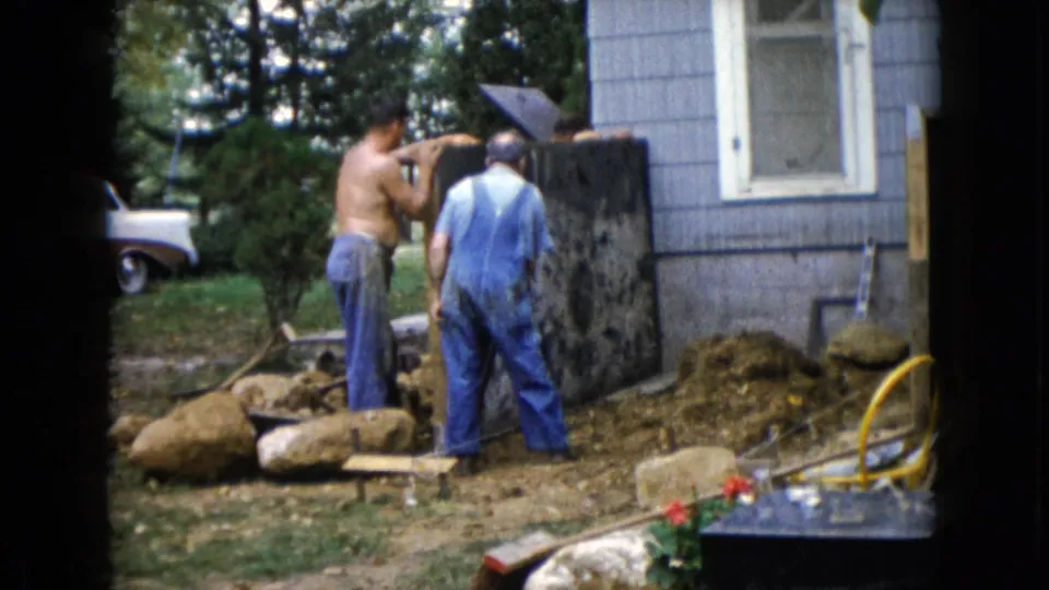 The men continue working around the dark slab beside the house, with rocks, dirt, trees, and part of a white car in the background.