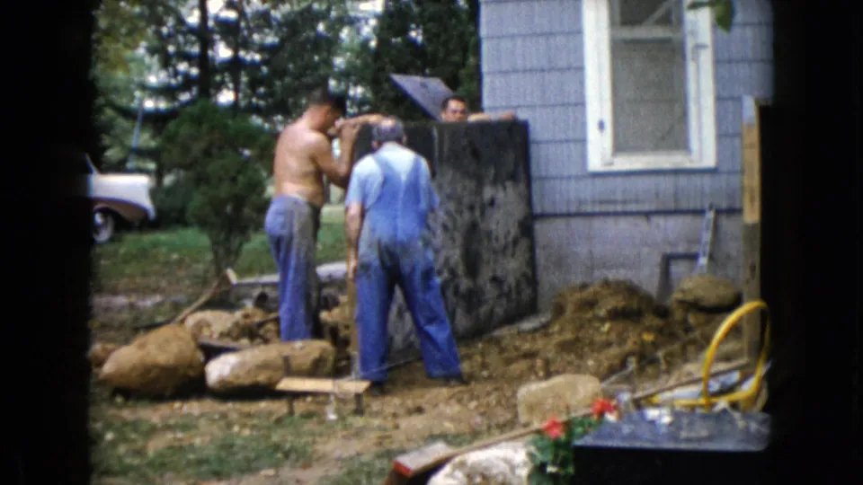 At the end of the clip, two men remain by the slab and a third visible face appears behind it near the blue-sided house.