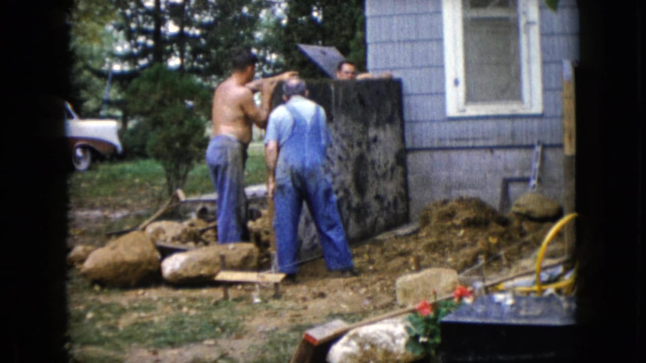 Three men work beside a blue-sided house with dirt, rocks, boards, and a partly visible white car nearby.