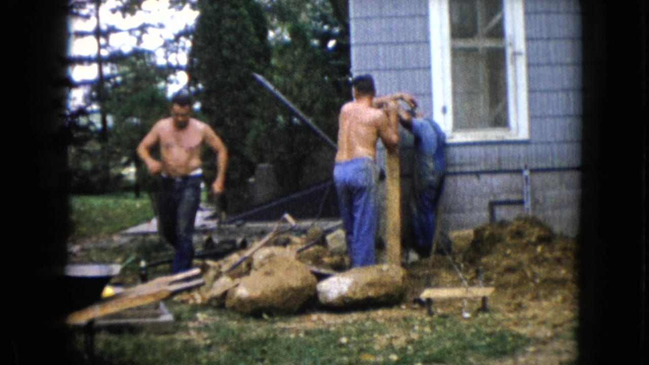 A 1960 Wauconda yard scene shows a shirtless man walking as two others handle a wooden post near the blue-sided house and scattered construction materials.