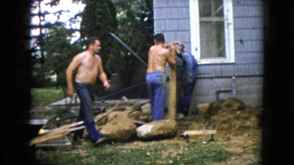 The home-movie frame shows informal 1960 construction activity in a Wauconda yard, with one man walking and two men positioned around a post near the house.