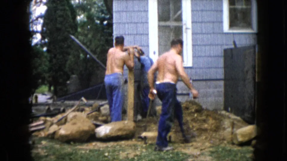 In the final 1960 Wauconda frame, two men remain at the post beside the blue house while a shirtless man walks across the construction area.